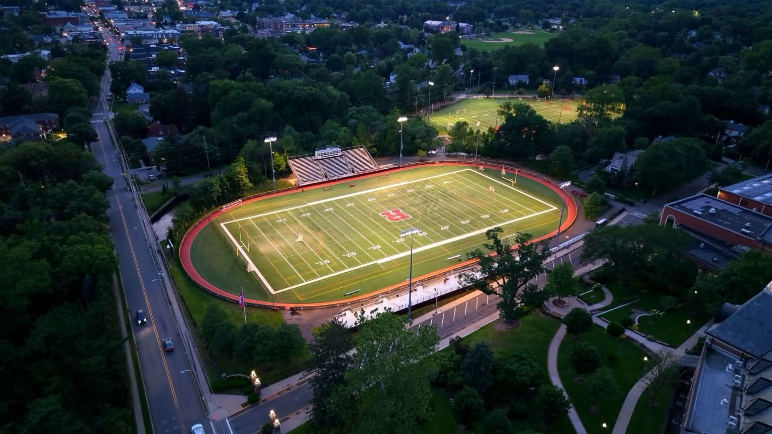 Dusk aerial of a New Jersey stadium and turf football field framed by a tree-lined town
